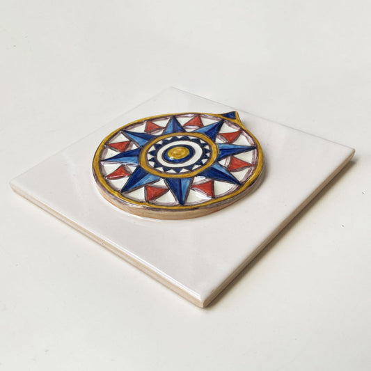 Azulejo quadrado em cerâmica vidrada branca exibindo uma rosa dos ventos geométrica em relevo pintada à mão em tons de azul, vermelho e amarelo, com acabamento tridimensional.