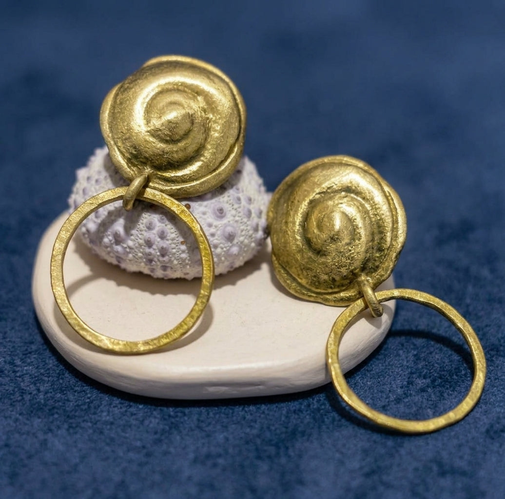 Brincos Concha em latão dourado com topo espiralado e argola pendente sobre suporte de cerâmica e fundo azul.