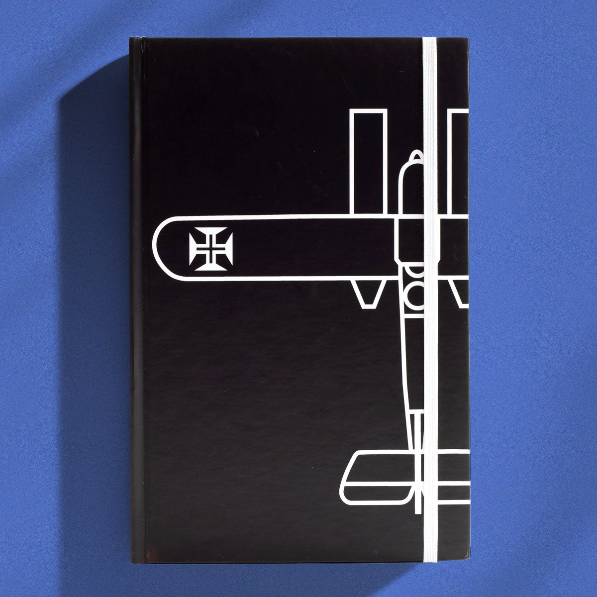Caderno de capa dura preta tamanho A5 com desenho minimalista em linhas brancas do Hidroavião Santa Cruz de Gago Coutinho e elástico de fecho vertical branco.