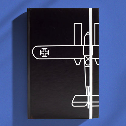 Caderno de capa dura preta tamanho A5 com desenho minimalista em linhas brancas do Hidroavião Santa Cruz de Gago Coutinho e elástico de fecho vertical branco.