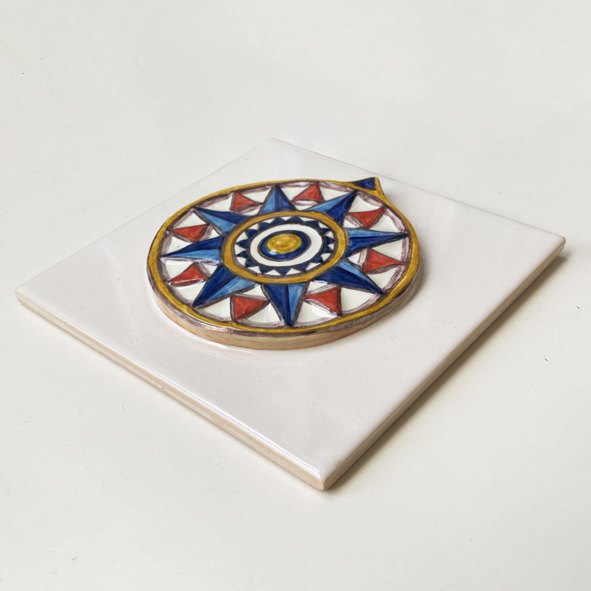 Azulejo quadrado em cerâmica vidrada branca exibindo uma rosa dos ventos geométrica em relevo pintada à mão em tons de azul, vermelho e amarelo, com acabamento tridimensional.