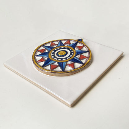 Azulejo quadrado em cerâmica vidrada branca exibindo uma rosa dos ventos geométrica em relevo pintada à mão em tons de azul, vermelho e amarelo, com acabamento tridimensional.
