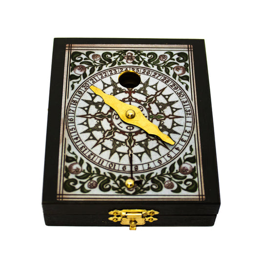 Bússola Kepler fechada na sua caixa de madeira retangular escura, destacando o fecho metálico dourado e o topo decorado com mostrador ornamental numérico e ponteiro.
