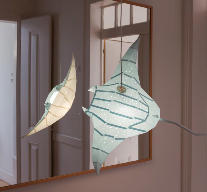 Candeeiro decorativo geométrico de papel em formato de raia marinha aceso sobre uma mesa, emitindo uma luz ambiente suave e realçando a silhueta poligonal do animal.
(EN: Geometric decorative paperlamp shaped like a marine stingray lit up on a desk, emitting a soft ambient glow and highlighting the animal's polygonal silhouette.)
