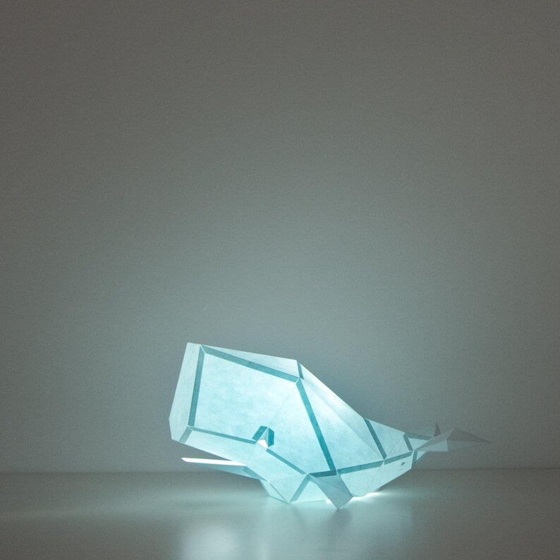 Candeeiro decorativo de papel azul suave em formato de baleia cachalote com design geométrico moderno aceso, emitindo uma luz suave sobre uma superfície plana.