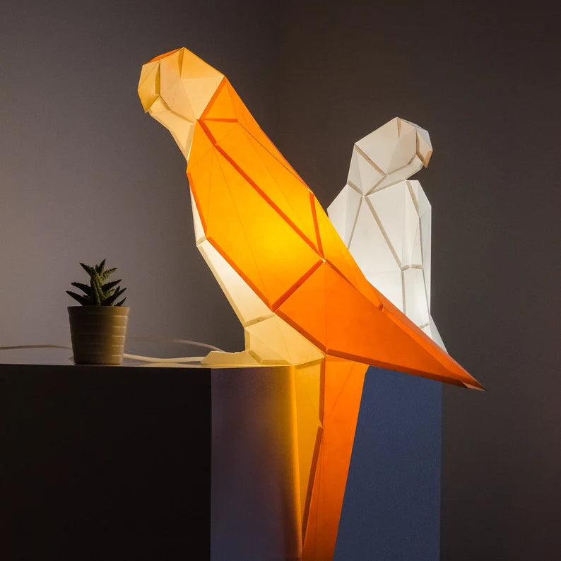 Candeeiro decorativo geométrico de papel em formato de papagaio tropical aceso sobre uma mesa, emitindo uma luz quente e realçando o design papercraft.