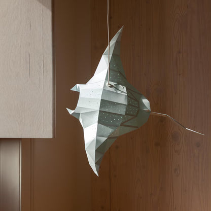 Candeeiro decorativo geométrico de papel em formato de raia marinha aceso sobre uma mesa, emitindo uma luz ambiente suave e realçando a silhueta poligonal do animal.
(EN: Geometric decorative paperlamp shaped like a marine stingray lit up on a desk, emitting a soft ambient glow and highlighting the animal's polygonal silhouette.)