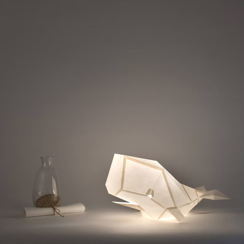 Candeeiro decorativo de papel de cor beige claro em formato de baleia cachalote com design geométrico moderno aceso, emitindo uma luz suave sobre uma superfície plana.
