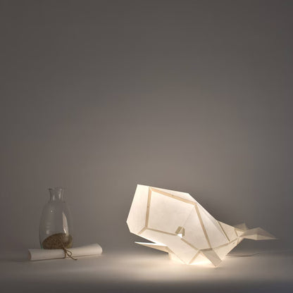 Candeeiro decorativo de papel de cor beige claro em formato de baleia cachalote com design geométrico moderno aceso, emitindo uma luz suave sobre uma superfície plana.